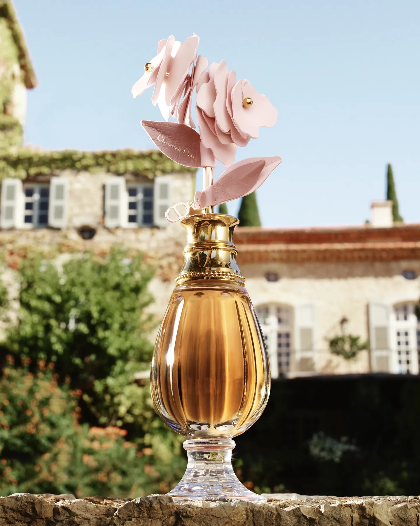 Flacon de l'édition limitée Parfums Christian Dior coiffé d'une rose en cuir, devant une bâtisse en pierre aux volets clairs