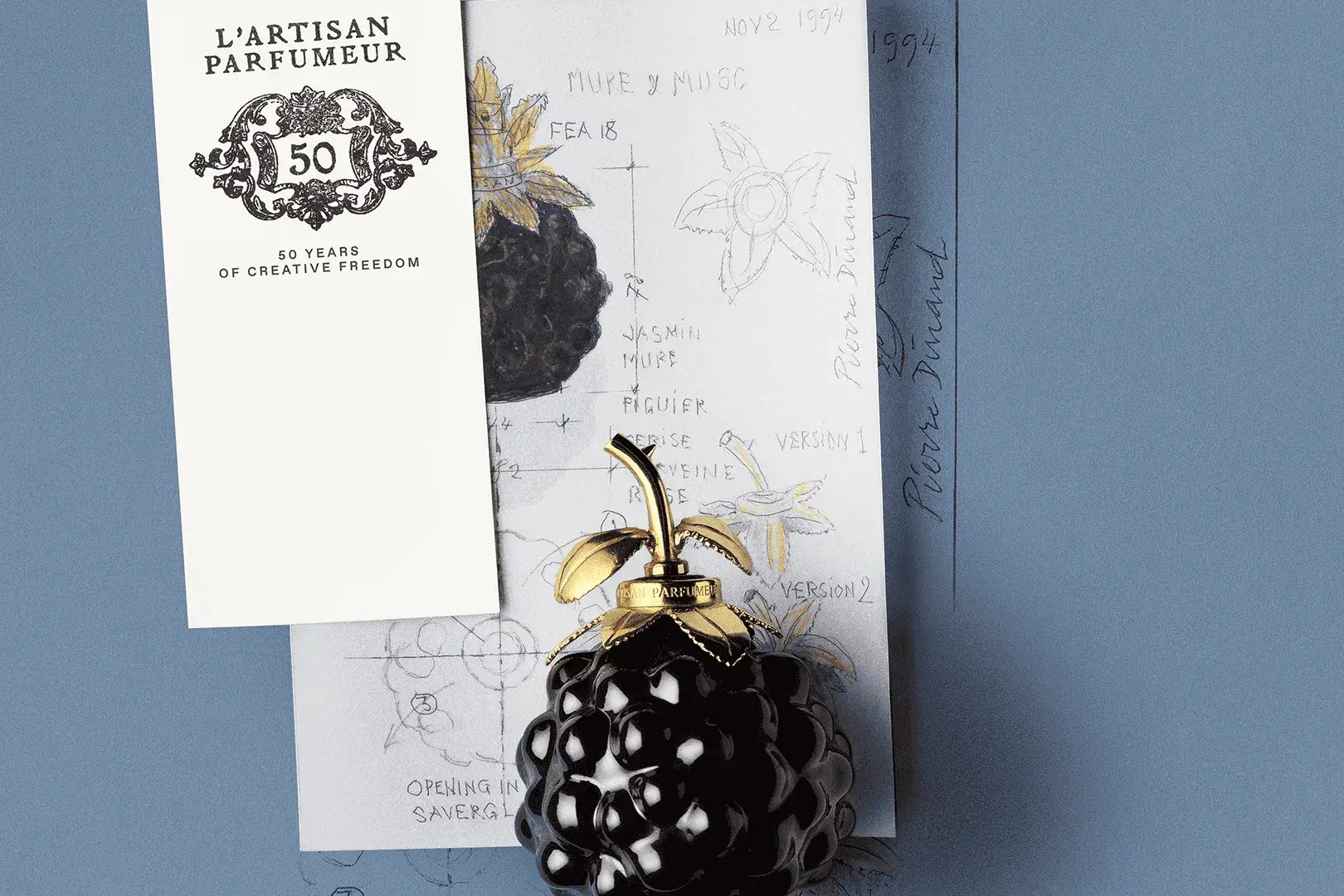 Carte des 50 ans de L'Artisan Parfumeur et flacon noir en forme de mûre à feuilles dorées sur fond bleu